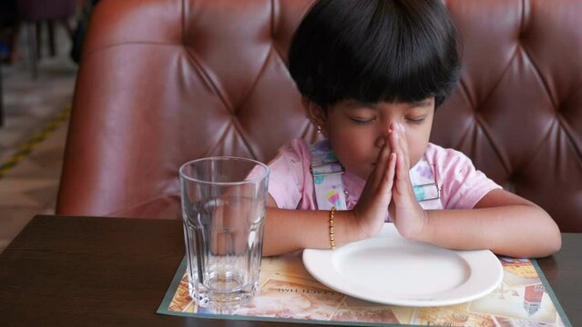 Kid praying before food in restaurant waiting for food Pink dress and plate and glass in front of her