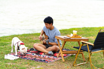 Asian man playing guitar during picnic with his French bulldog at pets friendly park. Domestic dog...