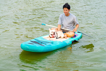 Asian man with his French bulldog paddle boarding in the lake during travel together at pets...