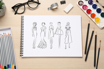 Sketches of different clothes in pad on wooden table. Fashion designer's desk with stationery, flat lay