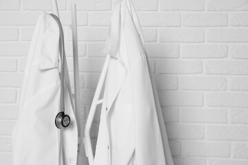 Medical uniforms and stethoscope hanging on rack near white brick wall. Space for text