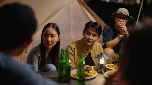 4K Group of Happy Asian people having dinner and talking together during camping in forest at night. Man and woman friends enjoy outdoor adventure lifestyle travel nature on summer holiday vacation.
