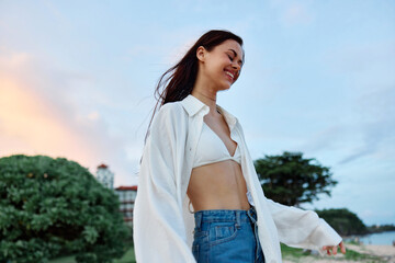 Brunette woman with long hair in a white shirt and jean shorts tan body abs and happiness fun smile with teeth on the beach and palm trees, vacation summer trip sunset sky
