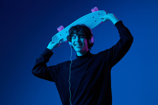 Teenage male with headphones listening to music and dancing and singing with skateboard in hand over his head, hipster lifestyle, blue background, neon light, style and trends, mixed light, copy space