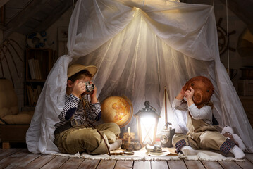 Obraz na płótnie Canvas Children play in the attic of their house. Imagine themselves as tourists, explorers. Look through a telescope, study a world map, play in a makeshift tourist tent. Dreaming of travel and adventure.