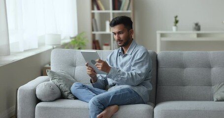 Smiling Indian guy spend free time sit on sofa holds digital tablet, scroll information, websurfing, check mail, texting message on device, enjoy new on-line game, rest at home use Generative AI