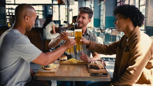 Group of multi-cultural male friends meeting in bar eating burger and fries and doing cheers with beer - shot in slow motion
