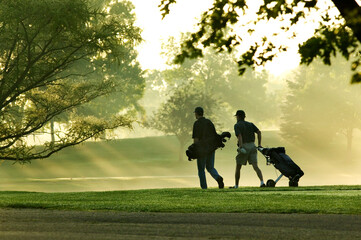 Two men golfers walking on a golf course with the sun streaking through the trees on a beautiful...