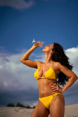 Attractive girl in bikini pouring liquid on to herself from wine glass, artistic image, clouds in background 