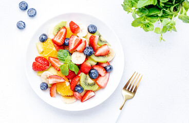 Fresh summer fruit and berry salad with strawberries, blueberries, banana, kiwi, orange, mint leaves and honey dressing, white table background, top view