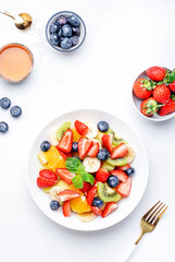 Fresh summer fruit and berry salad with strawberries, blueberries, banana, kiwi, orange, mint leaves and honey dressing, white table background, top view