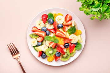 Summer healthy fruit and berry salad with strawberries, blueberries, banana, kiwi, orange and mint leaves, pink background, top view