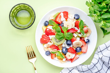 Summer fruit and berry salad with strawberries, blueberries, banana, soft cheese and mint leaves, green background, top view