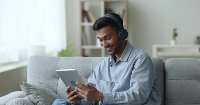 Handsome Indian guy spend pastime having video call conversation through app, sit on sofa using digital tablet, share news, talk to family living abroad, enjoy webchat virtual meeting Generative AI
