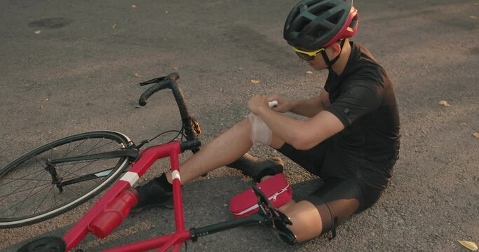 young athlete applying bandage onto his knee near bicycle outdoors in the evening. First aid after injury trauma slow motion