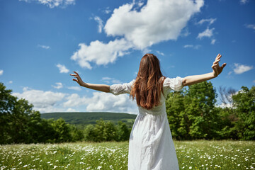 a woman stands with her back to the camera in a light dress in a chamomile field with her hands raised above her head
