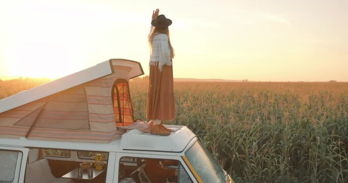 Beautiful careless relaxed happy woman dancing on the top of retro yellow camper van, enjoying beauty of landscape, corn field amazing nature in summer evening Freedom Happiness Lifestyle Slow motion