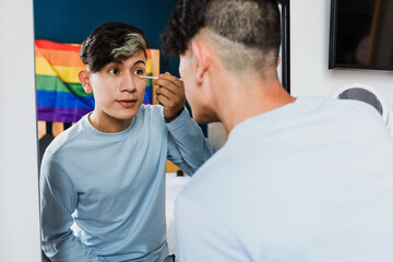 Young latin homosexual man applying makeup with mirror in bedroom at home in Mexico, Hispanic homosexual and lgbtq community in Latin America