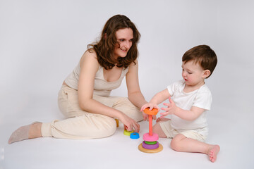 Happy baby with mother play educational toys on studio white background. Portrait of a smiling child with mom and playing while sitting on the floor. Kid about two years old (one year nine months)