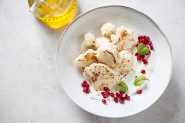 Plate with roasted cauliflower florets, cream-cheese and pomegranate, horizontal shot on a light-beige stone background, elevated view