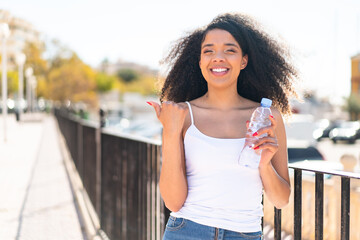 Young African American woman with a bottle of water at outdoors pointing to the side to present a product