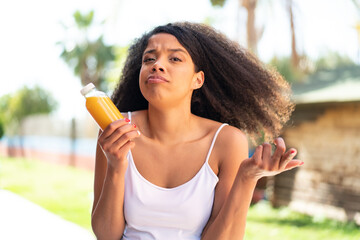 Young African American woman holding an orange juice at outdoors making doubts gesture while lifting the shoulders