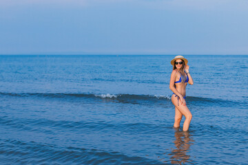 girl in a blue swimsuit, sunglasses and boater poses in seawater