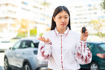 Young Chinese woman holding car keys at outdoors making doubts gesture while lifting the shoulders