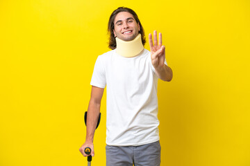 Young handsome man wearing neck brace and crutches isolated on yellow background happy and counting three with fingers