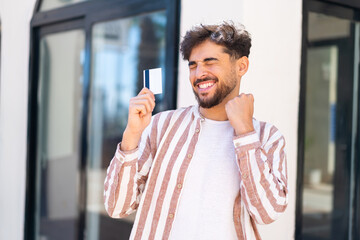 Handsome Arab man at outdoors holding a credit card and celebrating a victory