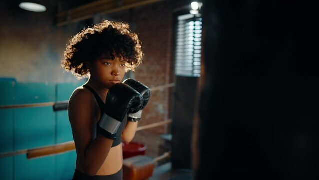Portrait of afro american woman boxer with curly hair boxing in gym on boxing ring. Young girl boxing training punches on punching bag. Professional sport, workout, train and fighter training concept.