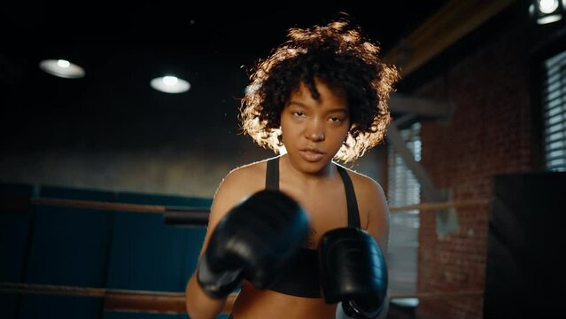 Afro american woman boxer in boxing gloves looks at camera stands on boxing ring and boxing. Sportswoman training in gym. Girl's power, woman train workout, sport fight, self-defense and feminism.