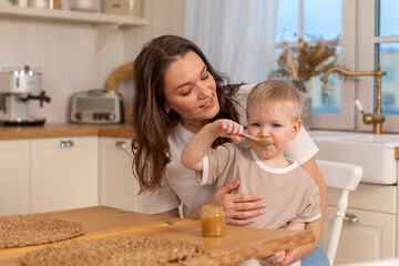 Obraz na płótnie Canvas Happy family at home. Mother feeding baby in kitchen. Little boy with messy funny face eats healthy food. Child learns eat by himself holding spoon. Woman mom giving food to kid son. Self feeding