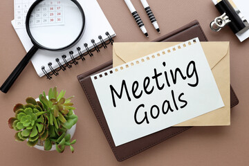 Office desk with text on a notebook. magnifying glass, pen, glasses, calendar and a small plant on a brown background. MEETING GOALS