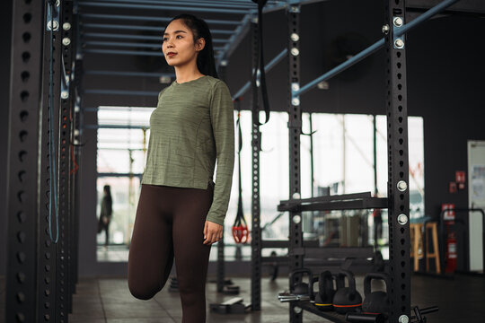 Strong asian woman streching body before exercise at crossfit gym. Athlete female wearing sportswear warm-up before workout on grey gym background with weight and dumbbell equipment.