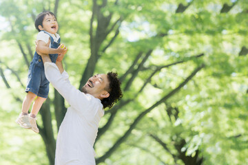 新緑の中高い高いをしてもらう子どもと幸せそうな家族　右にコピースペースあり