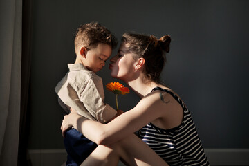 Real moment stylish Child Son gives mother gerbera flower for happy mother's day against dark wall. Authentic home life, hard light, sunny