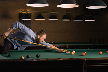 concentrated caucasian man in casual clothes playing billiards. He leaned over the table and makes a blow with a cue on the ball.