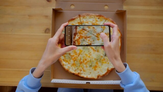 Top view - Female hands in blue sweatshirt use smartphone, take photo for social networks appetizing fresh cooked italian Quattro Formaggi Pizza in delivery box on wooden background. Generative AI