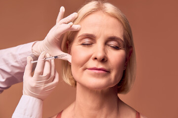 Aesthetic medicine.Portrait of an adult woman with closed eyes on a beige background. Hands in white gloves and a medical gown make an injection in the cheekbone. - obrazy, fototapety, plakaty