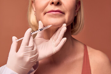 Cropped image of a woman getting an injection in her chin on a beige background. Hands in white...