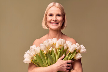 Portrait of a beautiful blonde middle-aged woman holding a bouquet of white tulips and smiling on an olive background.Women's beauty and spring.