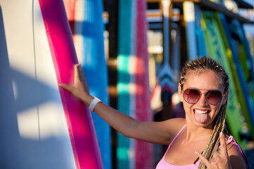 Fototapeta na wymiar Close-up of a tanned young woman in sunglasses with a rocker gesture standing at a sports equipment on the beach. Cool pretty girl imagining on camera Generative AI