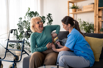Obraz premium Young dedicated nurse giving insulin injection to a diabetic female patient during home visit. Satisfied smiling senior woman receiving her daily therapy for blood sugar treatment at home.