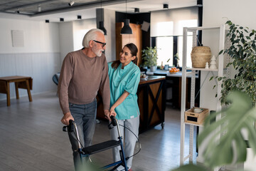 Kind supportive female physiotherapist helping elderly male patient with glasses to walk with...