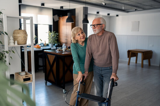 Blond Senior Woman Taking Care Of Her Husband With Physical Disability, Helping Him To Walk And Use Mobility Walker At Home. Senior Man Using Mobility Walker, Walking With His Wife Inside Apartment.