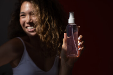 Smiling woman with lush curly hair and freckles on her face holds a bottle of cosmetic spray.