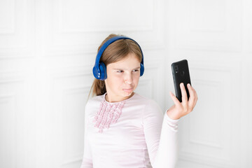 Naklejka premium Cheerful school girl wearing headphones, listening to music and taking a selfie on her smartphone in the class