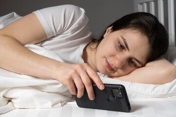 A woman lies in bed and reads the news or watches something on a smartphone before going to bed, gadgets in bed