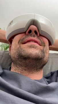 Young happy man using face and eye area massager. Relaxed man with closed eyes uses a massager for swelling and bruising around the eyes. The concept of male care and beauty treatments at home